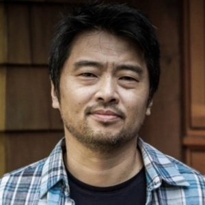Paul Bae stands in front of a shingled wall in a plaid shirt, looking at the camera.