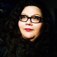 Andrea looks into the distance, wearing a black sweater and black rimmed glasses against a black background.