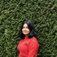 Hina wears a red sweater and stands in front of a hedge