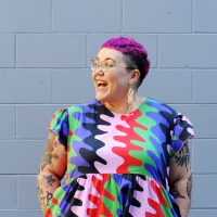 Image of Hannah McGregor in a brightly coloured dress, with pink short hair. She is smiling widely and looking off to her right.