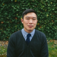 Eugene stands in front of a hedge and looks at the camera. He is wearing a dark sweater and tie, with a button up shirt underneath.