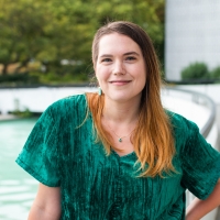 Grace stands smiling, wearing a green velvet top and black jeans.