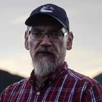 Chief Patrick Michell stands in front of a blurred mountain backdrop. He wears a baseball cap and a plaid shirt.